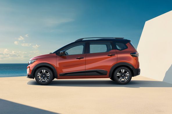 Red-orange compact SUV parked on a pale concrete surface by the sea, with a blue sky and a white angular wall in the background.