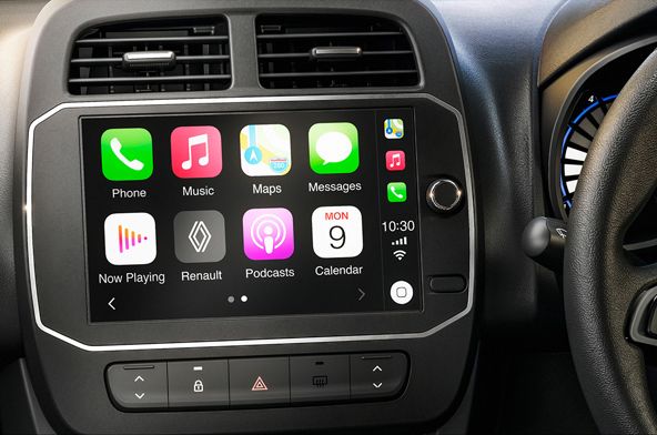 Car dashboard with a touchscreen showing Apple CarPlay icons (Phone, Music, Maps, Messages, Now Playing, Renault, Podcasts, Calendar) and physical controls below the screen, steering wheel on the right.